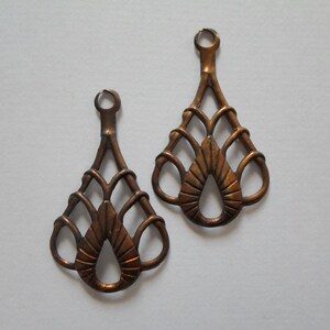May include: Two bronze-colored metal filigree teardrop-shaped charms. Each charm has a loop at the top for hanging and a decorative openwork design featuring a central teardrop shape and looped details.