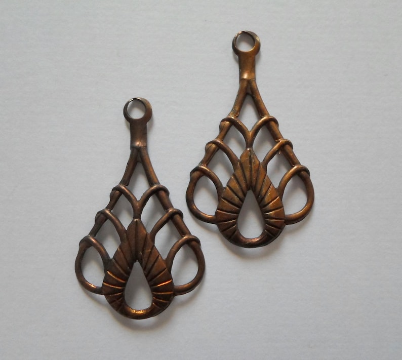 May include: Two bronze-colored metal filigree teardrop-shaped charms. Each charm has a loop at the top for hanging and a decorative openwork design featuring a central teardrop shape and looped details.