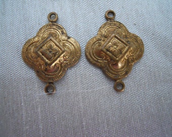 May include: Two antique gold-colored metal connectors. Each connector has a quatrefoil shape with a central diamond-shaped design featuring a floral element. The connectors have small loops at the top and bottom for attachment.