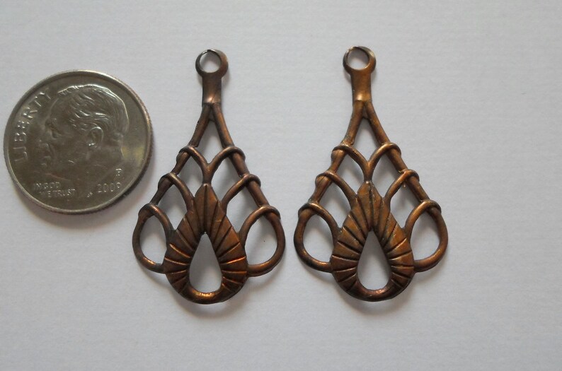 May include: Two copper-colored, openwork teardrop-shaped pendants with a small loop at the top. The design features a central teardrop shape with radiating lines and a woven pattern. A US quarter is next to the pendants.
