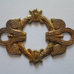 May include: Ornate, antique-style brass decorative hardware. The piece features a central open circle surrounded by detailed floral and scrollwork designs. The hardware has a warm, aged gold finish, suggesting vintage charm.