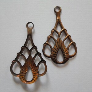 May include: Two ornate, bronze-colored metal pendants with a teardrop shape and intricate openwork design. Each pendant has a small loop at the top for hanging. The pendants have a vintage or antique aesthetic.