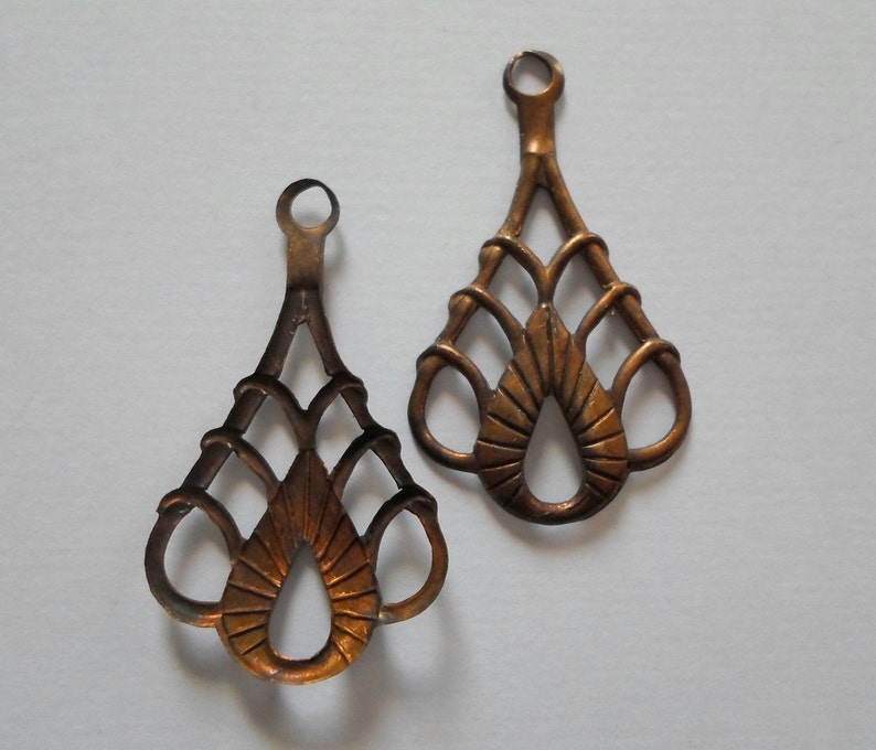 May include: Two ornate, bronze-colored metal pendants with a teardrop shape and intricate openwork design. Each pendant has a small loop at the top for hanging. The pendants have a vintage or antique aesthetic.