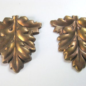 May include: Two gold-toned metal leaf-shaped embellishments with a raised, textured design. The leaves are detailed with a central vein and smaller leaves.