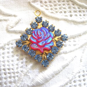 May include: A gold-toned pendant with a central rose design in shades of pink and blue, surrounded by light blue rhinestones. The pendant has a small loop at the top for hanging. The background is a white, textured fabric.