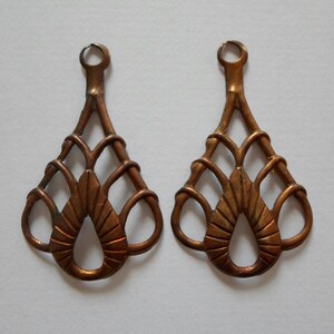 May include: Two ornate, copper-colored metal earring components. Each component features a looped top, a slender stem, and a decorative, openwork design with a teardrop-shaped central element and interwoven lines.