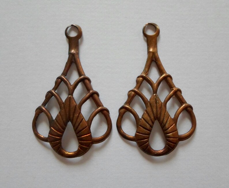May include: Two ornate, copper-colored metal earring components. Each component features a looped top, a slender stem, and a decorative, openwork design with a teardrop-shaped central element and interwoven lines.