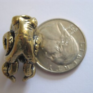 May include: A gold-colored metal frog charm with detailed sculpting. The charm is about the size of a US quarter.