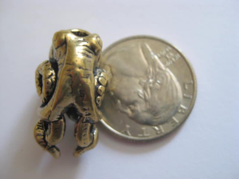 May include: A gold-colored metal frog charm with detailed sculpting. The charm is about the size of a US quarter.