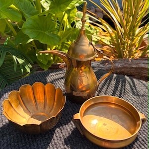 May include: A polished brass coffee pot with a tall, pointed lid and a long, curved spout, accompanied by a scalloped-edge bowl and a round, two-handled bowl. The coffee pot features an engraved palm tree design and a small rectangular label.