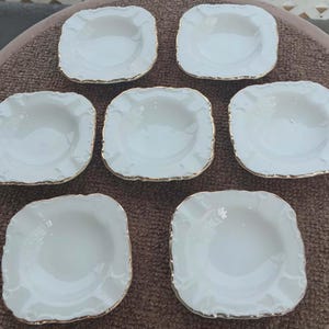 May include: Seven white, square-shaped dishes with scalloped edges and gold trim are arranged on a brown surface. The dishes have a shallow, bowl-like design, suitable for serving small portions.