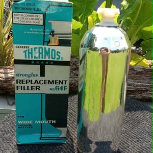 May include: A silver-colored glass vacuum bottle replacement filler with a white plastic stopper, standing next to its original blue and white cardboard box labeled Genuine Thermos Brand Stronglas Replacement Filler No. 64F for wide mouth bottles.