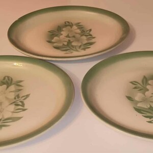 May include: Three oval-shaped plates with a white floral design and a green border. The plates are arranged on a white surface. The floral design features white flowers and green leaves.