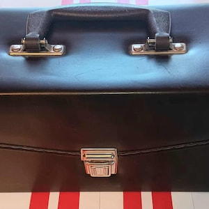 May include: A dark brown, rectangular briefcase with a handle and a metal clasp. The briefcase has a stitched design and metal accents on the handle attachments. The case is closed and appears to be made of leather or a leather-like material.