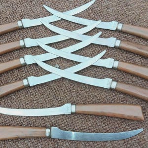 May include: Set of seven steak knives with curved stainless steel blades and brown wooden handles. The knives are arranged in a pattern, with some crossed over each other. The handles have a light brown color and a smooth finish.