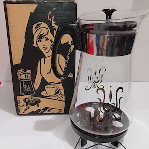 May include: A vintage glass coffee pot with a black handle and a black base. The pot has a decorative design on the side and a black lid. The pot is sitting on a black stand with three legs.