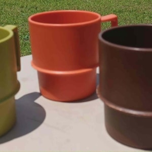 May include: Four vintage plastic mugs in shades of green, orange, and brown. Each mug has a handle and a unique stacked design. The mugs are arranged on a light-colored surface with a green background.
