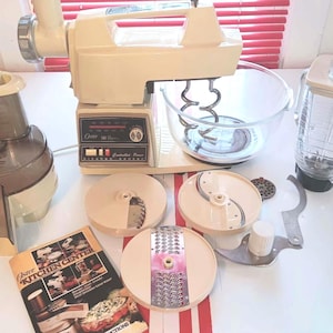 May include: An Oster Kitchen Center appliance set in cream and brown. Includes a mixer with a glass bowl, a blender, a juicer, and various slicing and grating attachments. A manual is also included.