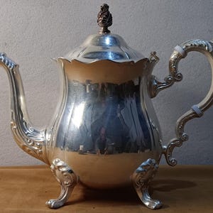 May include: A silver-colored teapot with ornate detailing. The teapot has a curved spout, a decorative handle, and rests on three small feet. The lid is topped with a brown, pinecone-shaped finial. The teapot is likely made of metal.