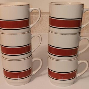 May include: Set of six white ceramic mugs with red, black, and gold stripes. The mugs are stacked in pairs.