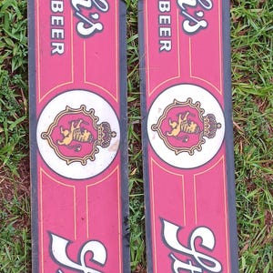 May include: Two long, rectangular vertical signs featuring a red background with black borders and gold trim. Each sign displays the white text Stroh's BEER twice, separated by a central white circle containing a gold lion crest with a crown.