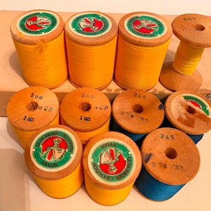 May include: A collection of wooden spools wound with yellow and blue nylon thread. Several spools feature green and red Gudebrod's Champion labels on the ends, while others are stamped with numbers like 209, 245, and 207, and weights in ounces.