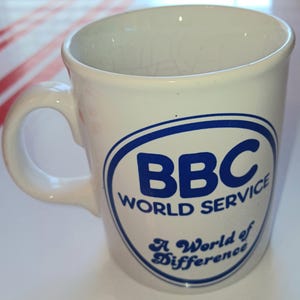 May include: White ceramic mug with a curved handle. The mug features the blue text "BBC WORLD SERVICE" inside a blue oval, with the text "A World of Difference" below. The mug is designed for hot beverages.