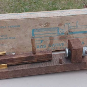 May include: A vintage wooden nutcracker with brass and metal components. The device is mounted on a wooden base, with a cylindrical brass piece and a box in the background. The box has text that reads "Texas Native".