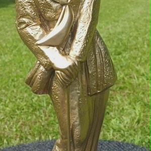 May include: A gold-colored statue of a golfer wearing a flat cap, a collared shirt with a neck scarf, and trousers. The figure stands on a square base, holding a golf club with both hands positioned in front of the body, featuring a textured finish.