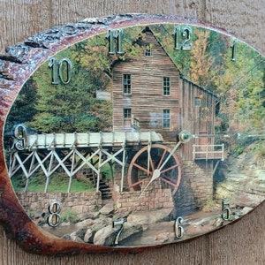 May include: A round wooden clock with a rustic wood grain finish. The clock face features a scenic image of a watermill with a waterfall and trees. The clock face has Roman numeral hour markers and a silver-colored hour and minute hand.