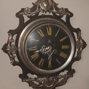 May include: An antique wall clock with a black face and gold Roman numerals. The clock is encased in a decorative silver-colored frame with ornate scrollwork. The clock hands are silver, and the overall design is vintage.