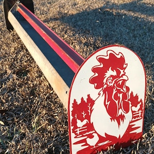 May include: A long, rectangular metal chicken feeder with a red and black interior. The end features a red and white rooster design with a rural landscape. The feeder is placed on a grassy surface.
