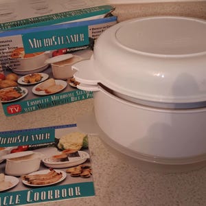 May include: A white two-tiered microwave steamer with a cookbook titled "Micro Miracle Cookbook" by Kitchenmate. The cookbook features images of food prepared in the steamer.