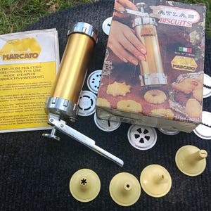 May include: A gold-colored metal biscuit press with a silver handle, accompanied by its original box labeled ATLAS BISCUITS and MARCATO, a multi-language instruction manual, and several interchangeable metal and plastic shaping discs and nozzles.