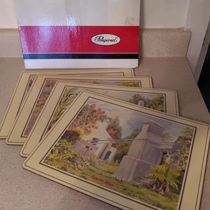 May include: Set of six beige placemats with a brown border. Each placemat features a watercolor painting of a house with a garden and the text "Balmetto House".