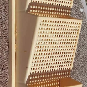 May include: A beige, three-tiered wall-mounted organizer with a rectangular design. Each tier features a grid-like pattern of small square holes. The organizer is made of a solid material and is designed to hold items.