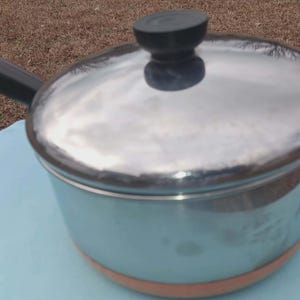 May include: A stainless steel saucepan with a lid and a black handle. The pot has a light blue exterior with a copper-colored base. The lid has a black knob. The pot is sitting on a light blue surface.