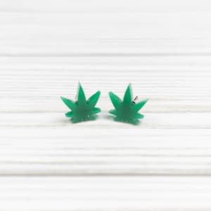 May include: A pair of green marijuana leaf stud earrings on a white wooden surface.
