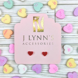 May include: A pair of red heart-shaped stud earrings displayed on a pink card with the text "J LYNN'S ACCESSORIES". The card is surrounded by candy hearts with various messages.