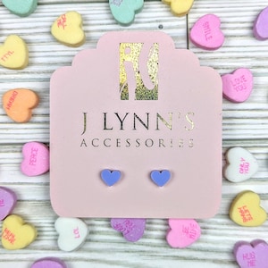 May include: A pair of lavender heart-shaped stud earrings displayed on a pink card with the text "J LYNN'S ACCESSORIES". The card is surrounded by small heart-shaped candies.