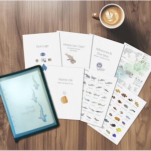 May include: A collection of dive-themed journals and guides, including a journal with the text "Dive Journal" and other guides with titles like "Dive Logs" and "Marine Life." A cup of coffee, a camera, and a watch are also on the wooden surface.