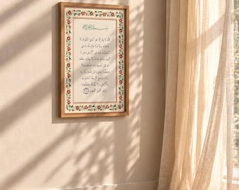 Op de afbeelding: Een rechthoekig wandkunstwerk met zwarte Arabische kalligrafie gecentreerd op een witte achtergrond, omringd door een decoratieve rand van rode en groene bloemmotieven, geplaatst in een lichtgekleurde houten lijst aan de muur.