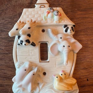 May include: A decorative white ceramic light switch cover featuring a house design with various animal figurines. The animals include giraffes, pandas, dolphins, ducks, and an elephant. The cover has a gold trim.