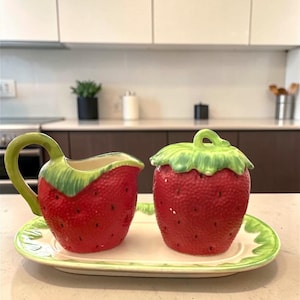 May include: A ceramic strawberry-themed creamer and sugar bowl set. The creamer has a green handle, and the sugar bowl has a green leafy top. Both are red with black speckles, sitting on a matching tray with a green border.
