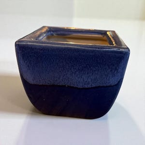 May include: A square ceramic planter with a two-tone blue glaze. The top half has a textured, lighter blue finish, while the bottom is a darker, smooth blue. The interior is a light tan color.