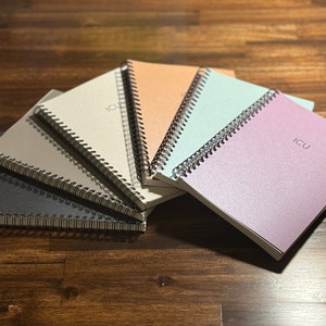 May include: A stack of six spiral bound notebooks in various colours, including black, grey, beige, light blue, peach, and lavender. The top notebook has the word "ICU" printed on the front.