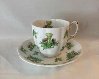 Peut inclure: Une tasse à thé et une soucoupe blanches avec un motif de chardon vert et violet. La tasse a une bordure dorée et une poignée dorée.