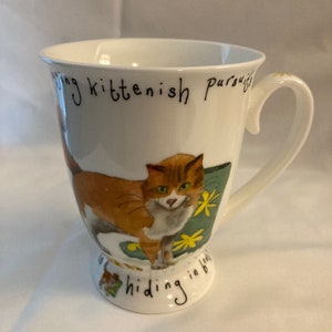 May include: White ceramic mug with a brown and white cat illustration. The mug has the text "Pursuing kittenish pursuits... hiding in bed" on the front.