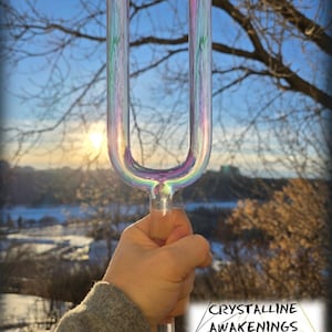 Puede incluir: Un diapasón de vidrio iridiscente sostenido en una mano contra un fondo de paisaje invernal con cielo azul y árboles. El diapasón está etiquetado como "Crystalline Awakenings, Lightwork & Soundhealing".