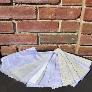 Op de afbeelding: Een verzameling witte stofstalen met verschillende texturen, waaronder glad, getextureerd en geëmbost. De stalen zijn in een waaier op een donker oppervlak gerangschikt.